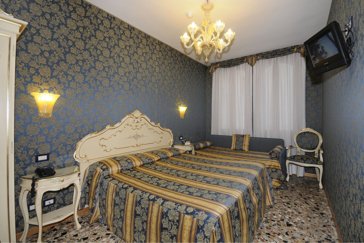 Une chambre avec un lit double, une petite table avec un téléphone, une chaise, un téléviseur fixé au mur et un lustre.