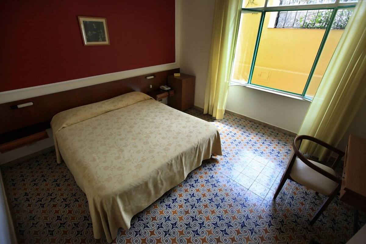 Una habitación con cama, una silla, una mesita, una ventana con cortinas y suelo estampado.