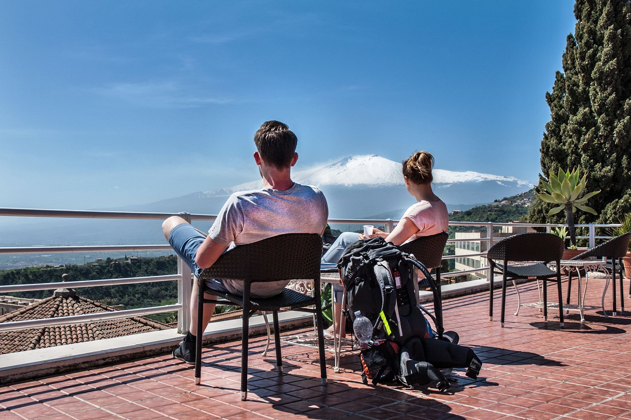Two people seated on outdoor chairs overlooking a scenic view with a mountain and a body of water in the background.