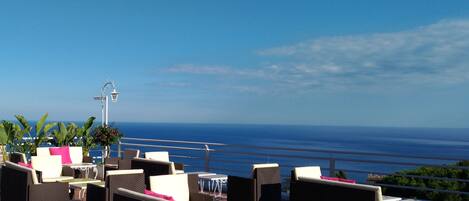 A terrace with wicker furniture and tables, overlooking a coastal view.