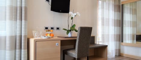 Habitación de hotel con un escritorio de madera, una silla estampada, un televisor de pantalla plana en la pared, una orquídea en maceta y una ventana con cortinas translúcidas.
