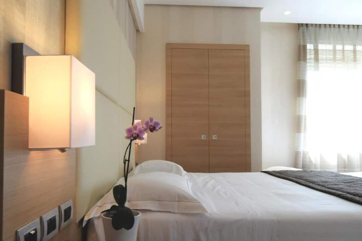 Una habitación de hotel con cama, mesita de noche, lámpara, armario y una orquídea en maceta.