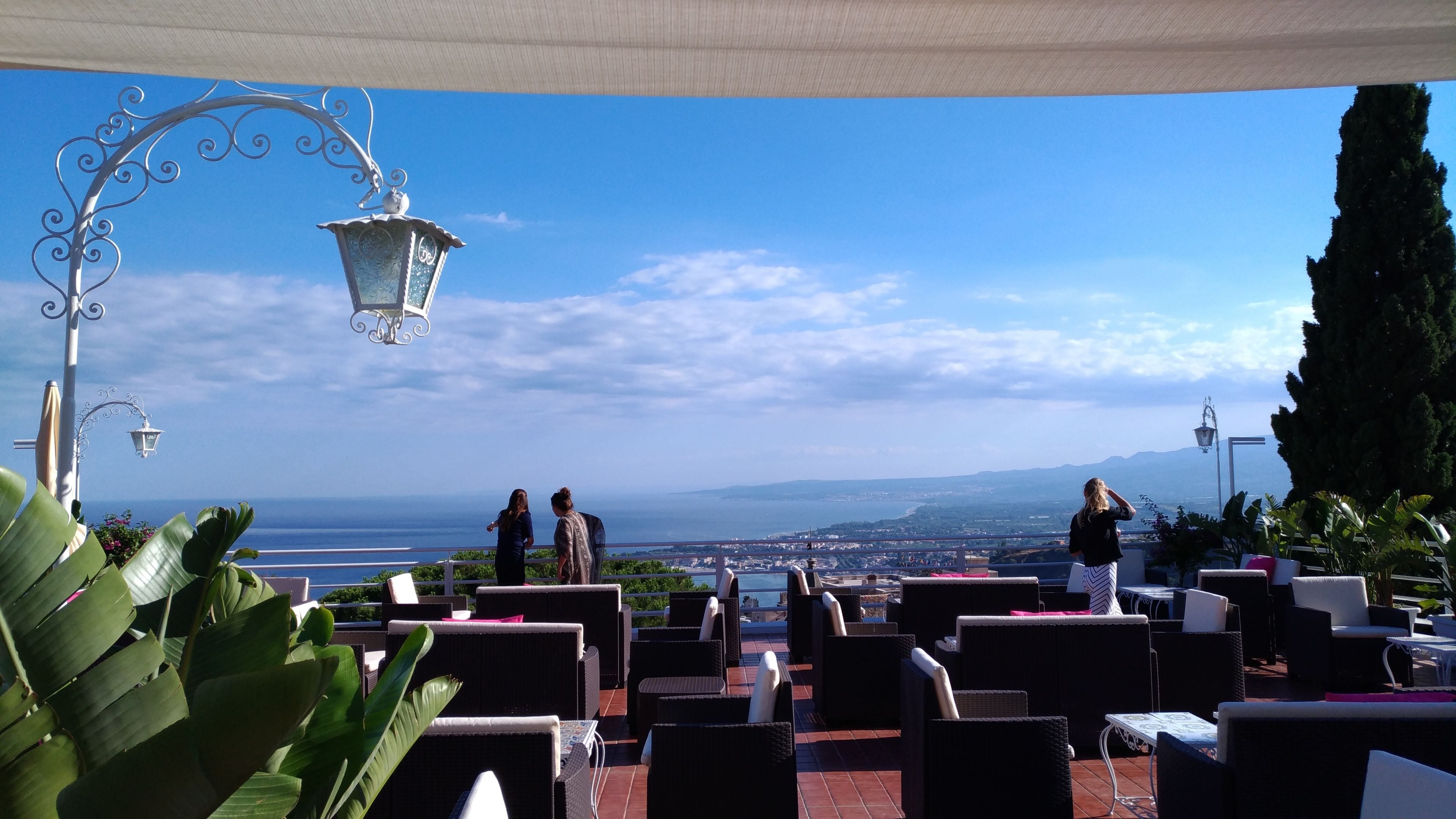 Un espacio al aire libre con sillas y mesas de mimbre, con vistas a un paisaje marino y una ciudad distante.