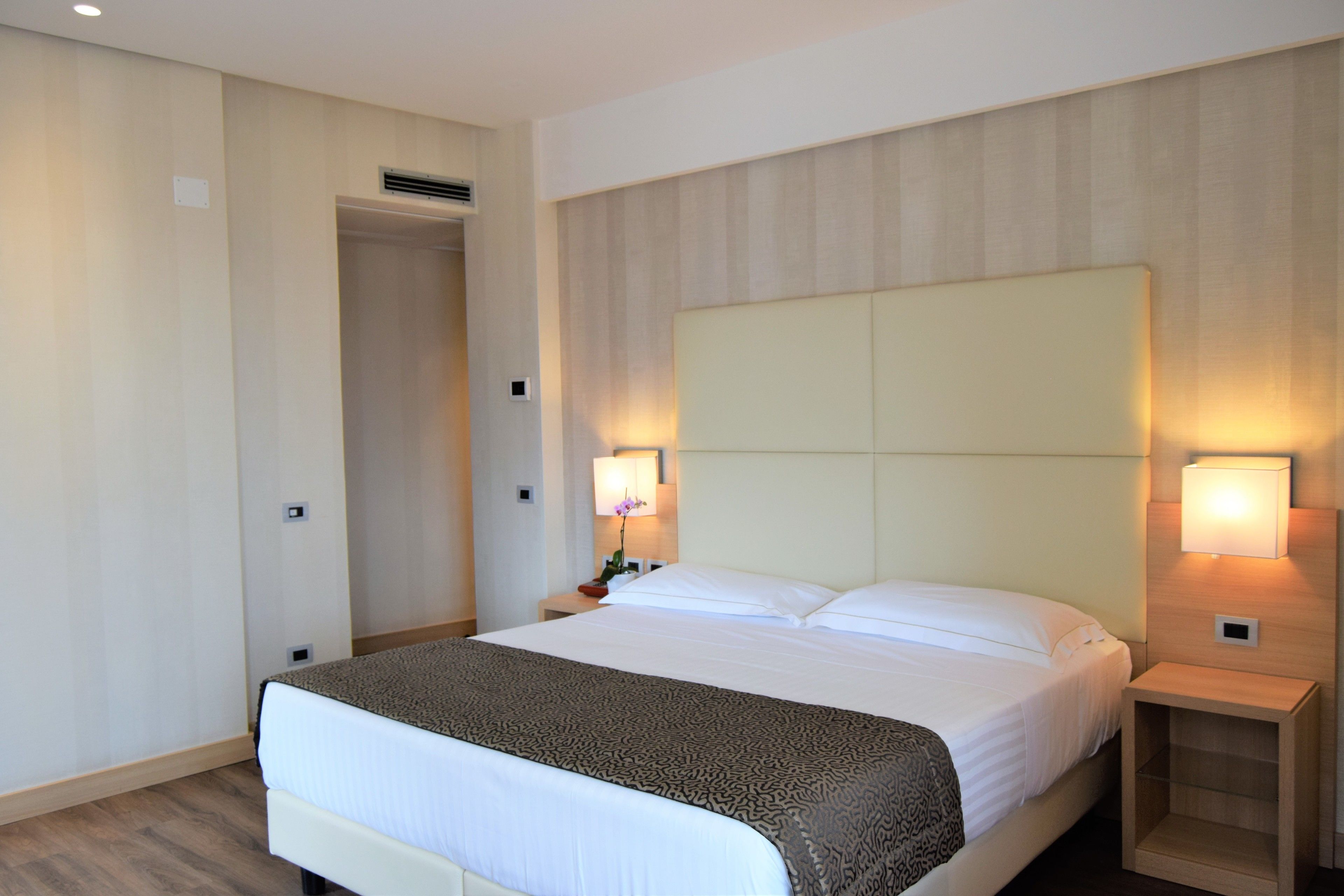 A hotel room with a large bed, two bedside lamps, a nightstand, and a door leading to another room.