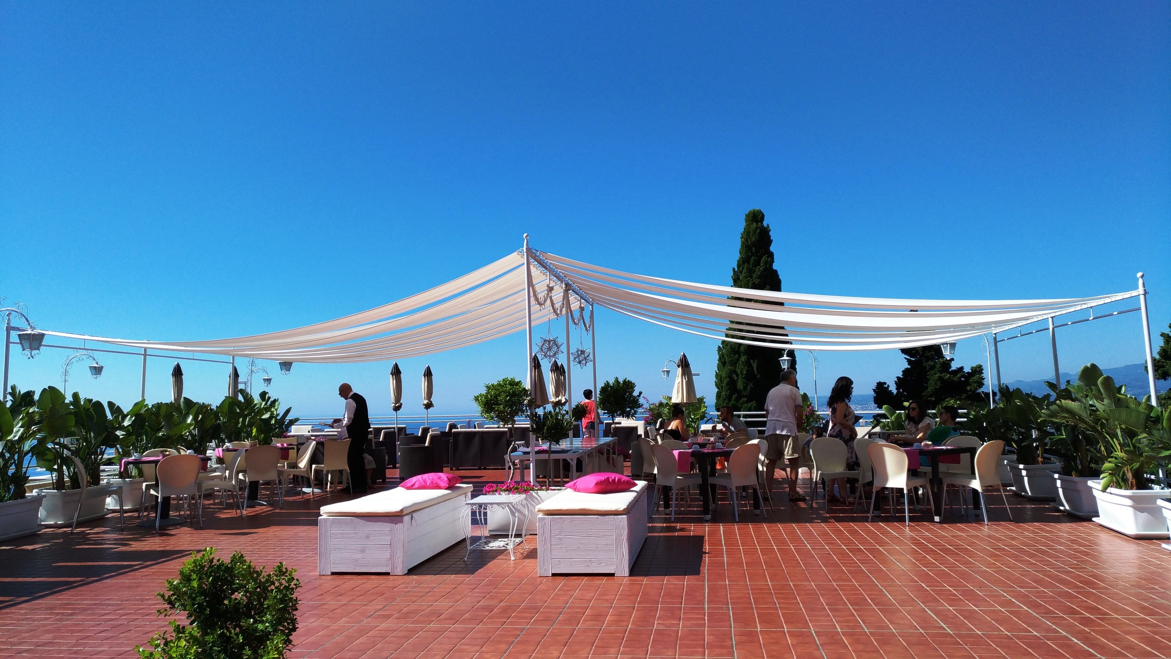 Terraza al aire libre con mesas, sillas, una toldilla y plantas en macetas.
