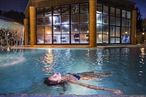 A swimming pool with clear water, a person swimming, and a building with large windows in the background. - Sardegna Termale Hotel & SPA (Sardara)