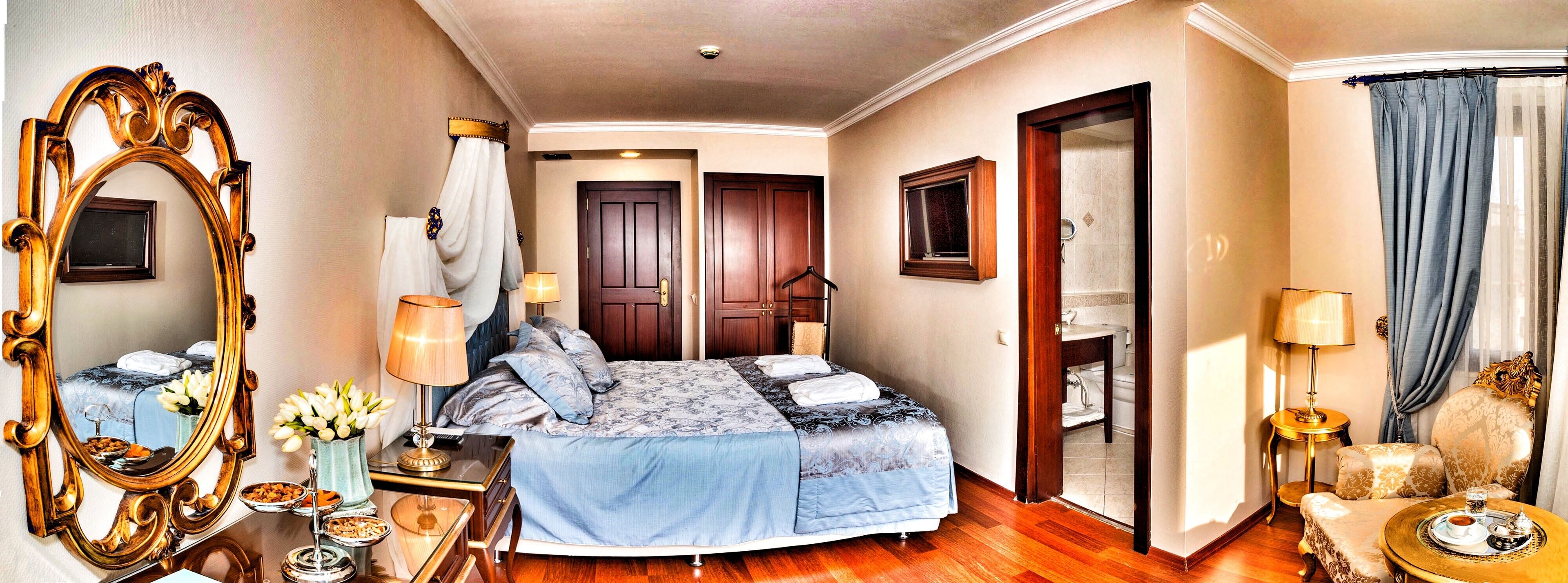 A hotel room with a large bed, a sitting area with a chair and table, a bathroom with a shower and toilet, and a mirror with a decorative frame.