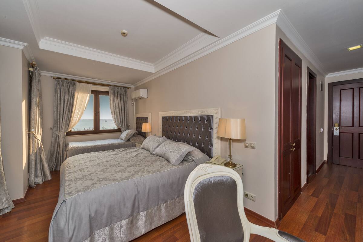 A hotel room with a large bed, a bedside table with a lamp, a chair, a window with curtains, and a door.