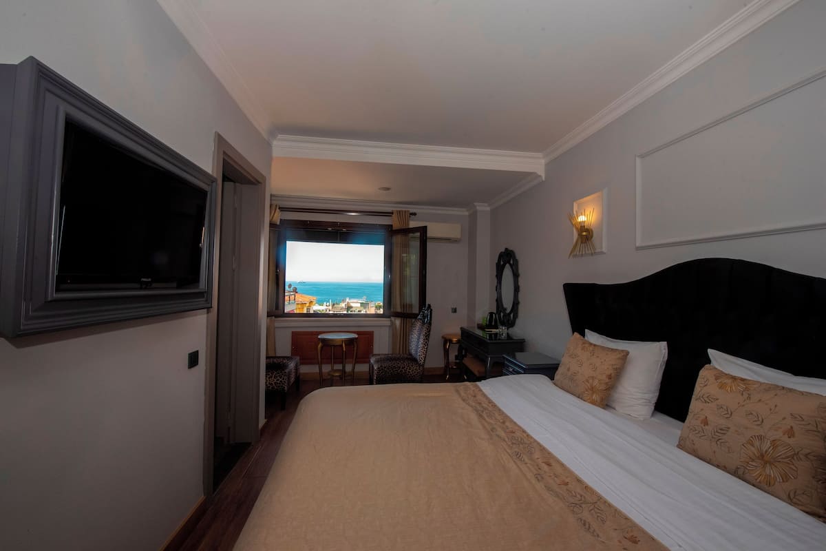 A hotel room with a bed, a TV, a desk with a chair, and a view of the sea.