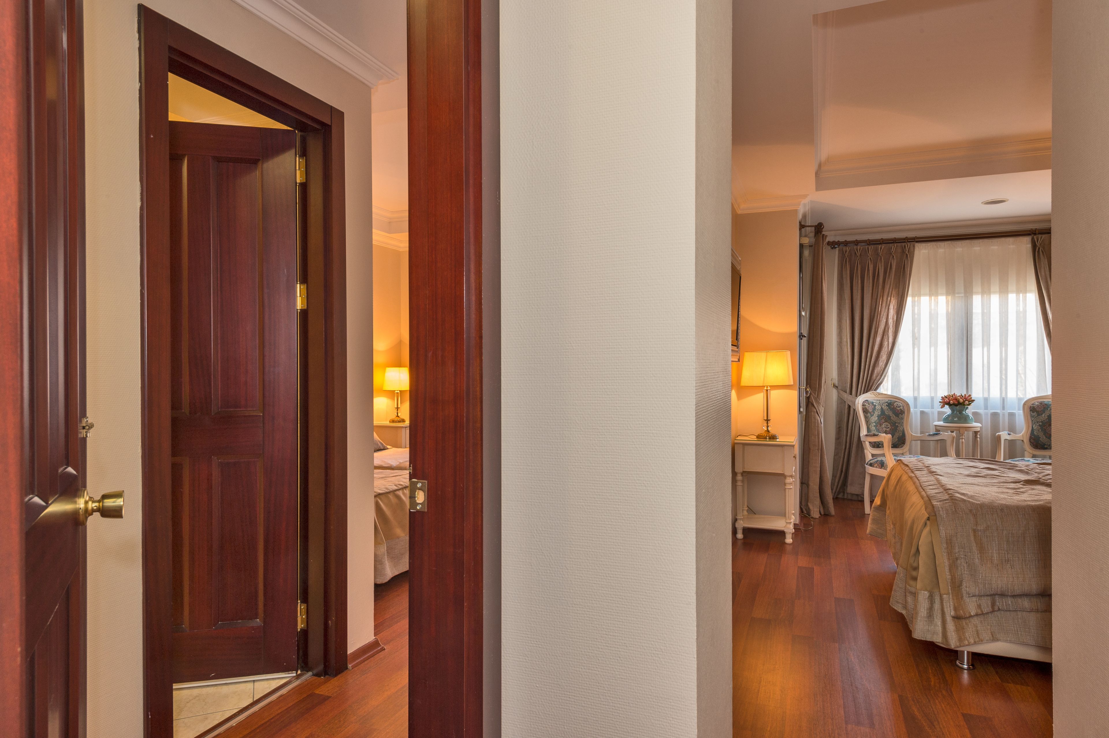 A hotel room with a wooden door, a bed with a beige bedspread, two bedside tables with lamps, a small table with a vase, and a window with curtains.