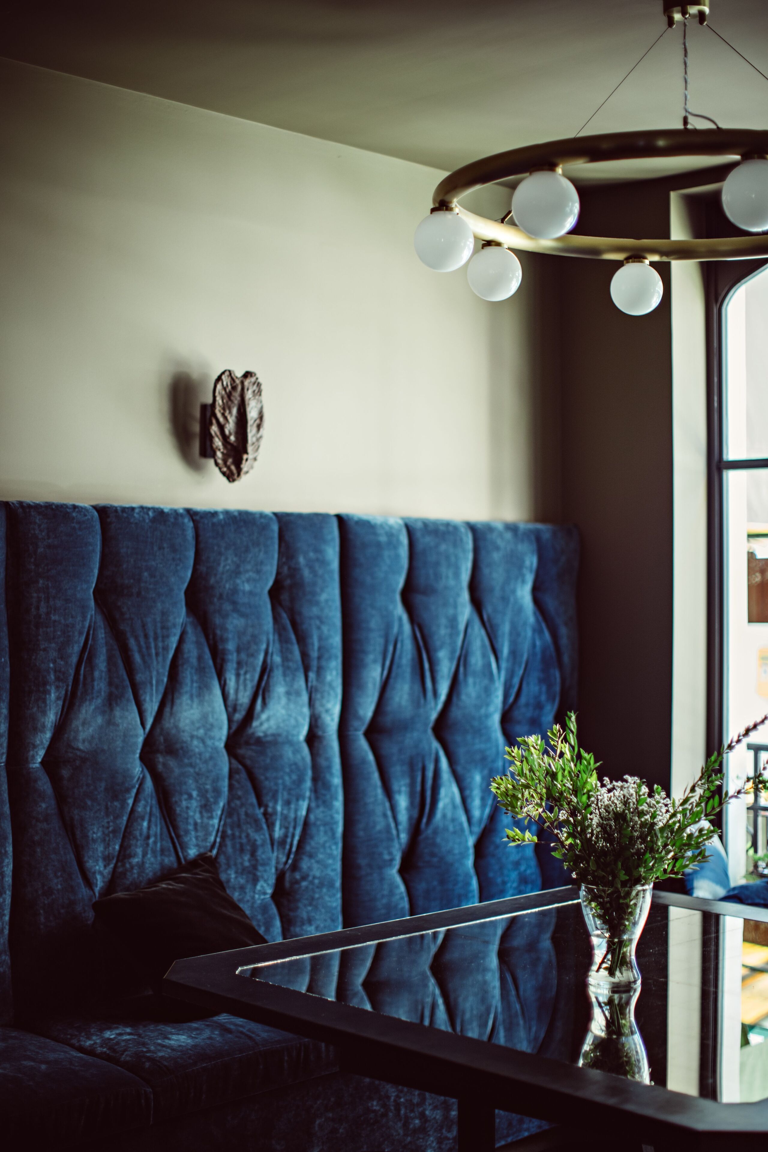 Een blauwe gestoffeerde bank met een gestuikte bekleding, een ronde tafel met een bloevas en een wandlamp met meerdere lampen.