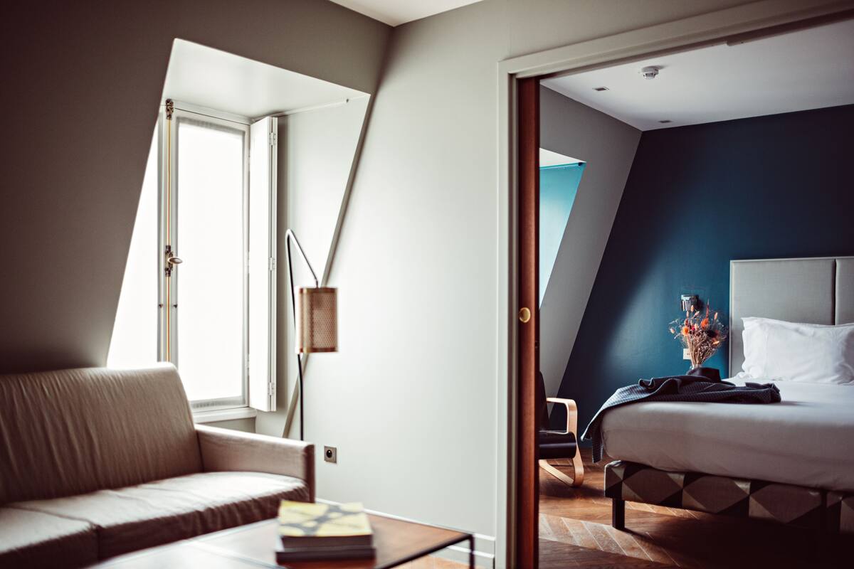 A modern hotel room with a bed, a sofa, a desk, and a large window.