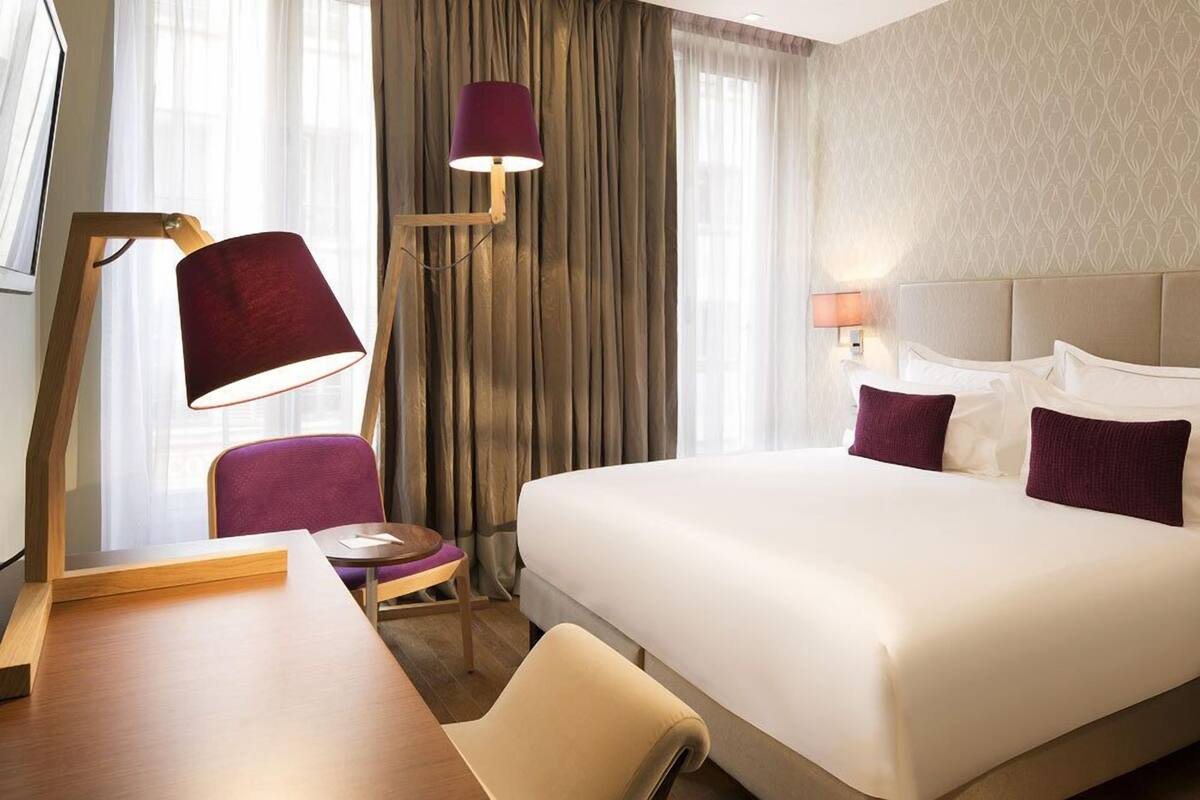 A hotel room with a large bed, a desk with a lamp, a chair, and a small table.