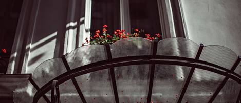 A metal outdoor chair with a mesh backrest, positioned in front of a building with windows and red flowers in a window box.