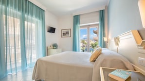 Una habitación de hotel con cama, una mesita de noche con un libro, televisor en la pared y una ventana con cortinas.