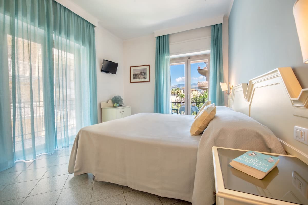 A hotel room with a bed, a bedside table with a book, a TV on the wall, and a window with curtains.
