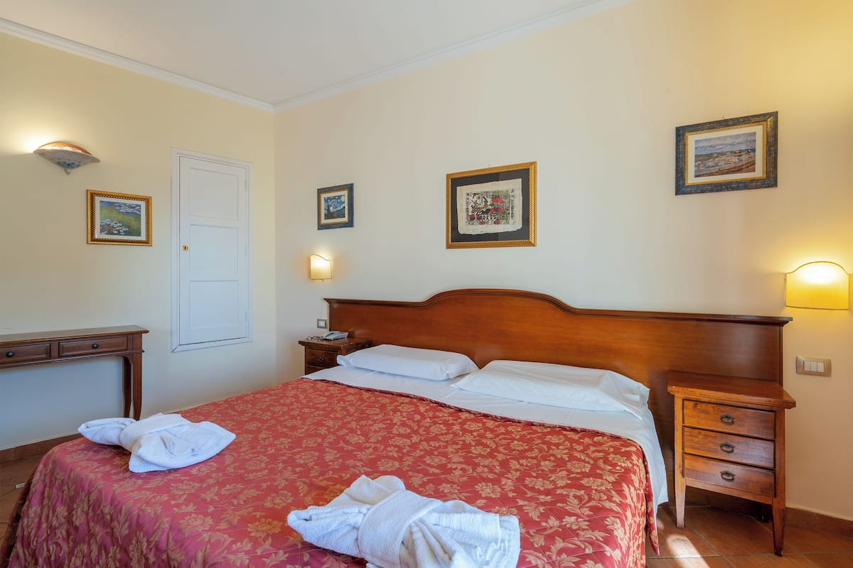 A neatly made bed with a red floral bedspread, a wooden nightstand with a lamp, and a wall with framed pictures.