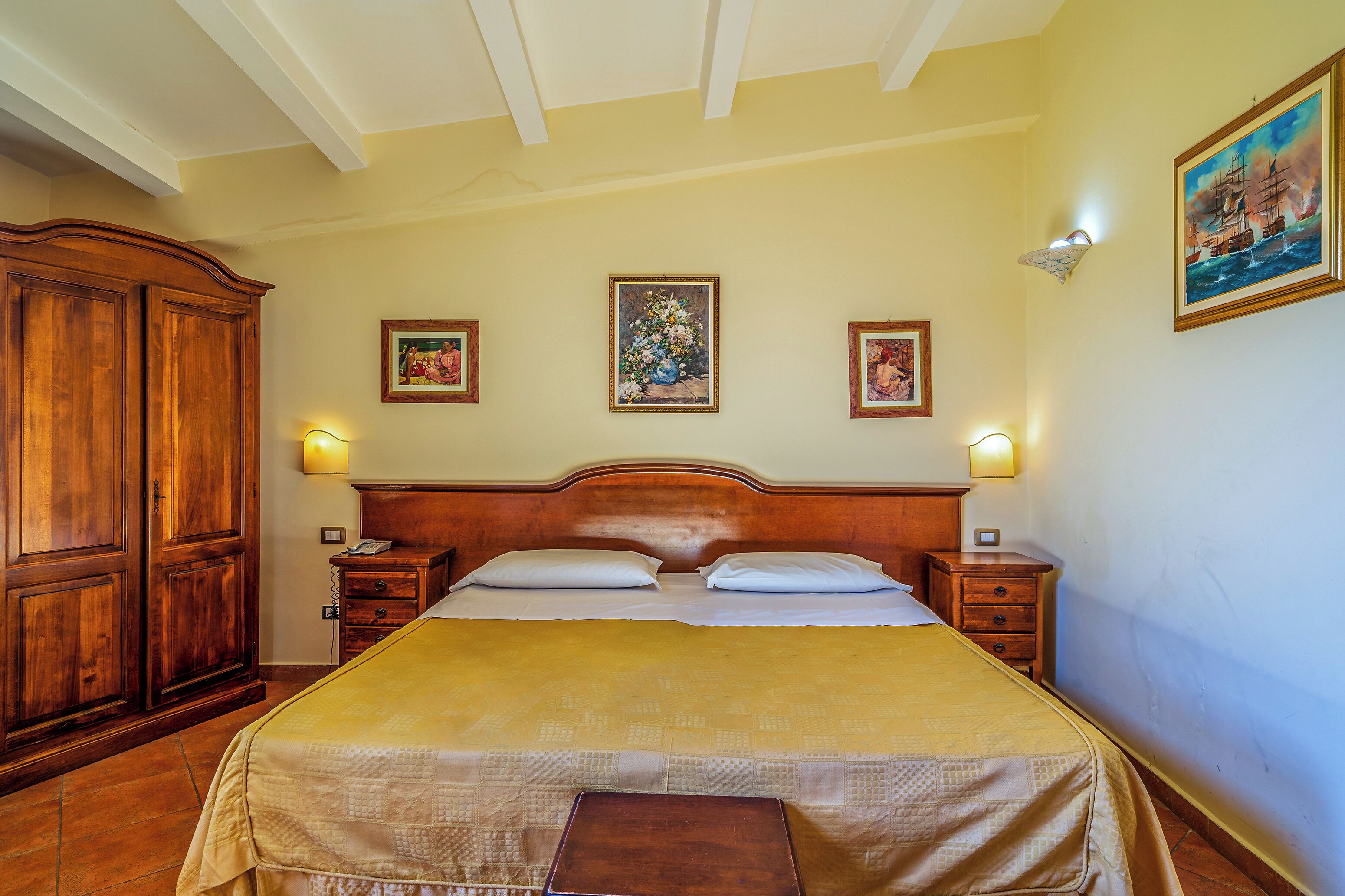 Habitación de hotel con una cama de madera, una mesita de noche y un armario. Hay tres cuadros en la pared.