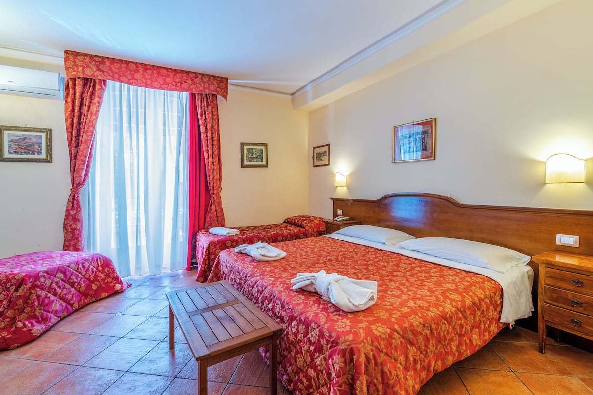 A hotel room with two beds, a wooden table, and a bench. There are two bedside tables with lamps and a wall-mounted light.