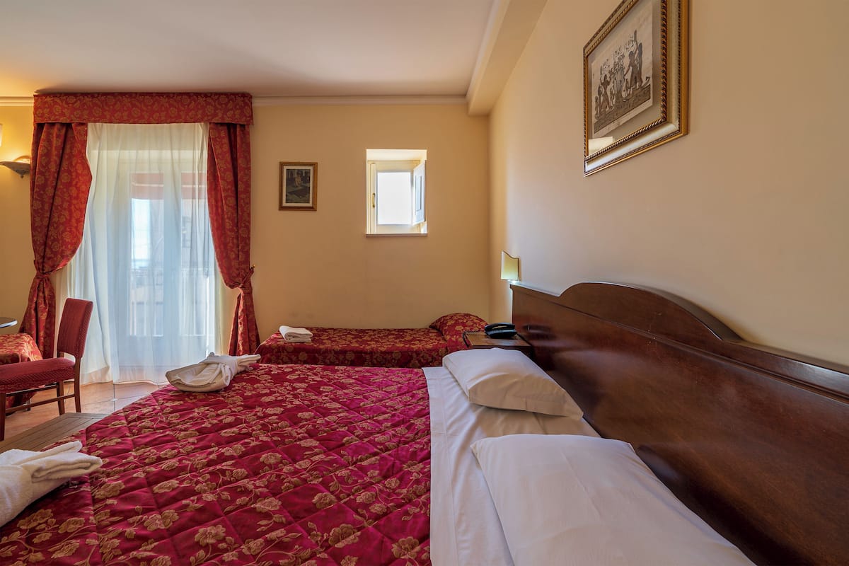 A hotel room with a large bed, two smaller beds, a chair, a desk, and a window with curtains.