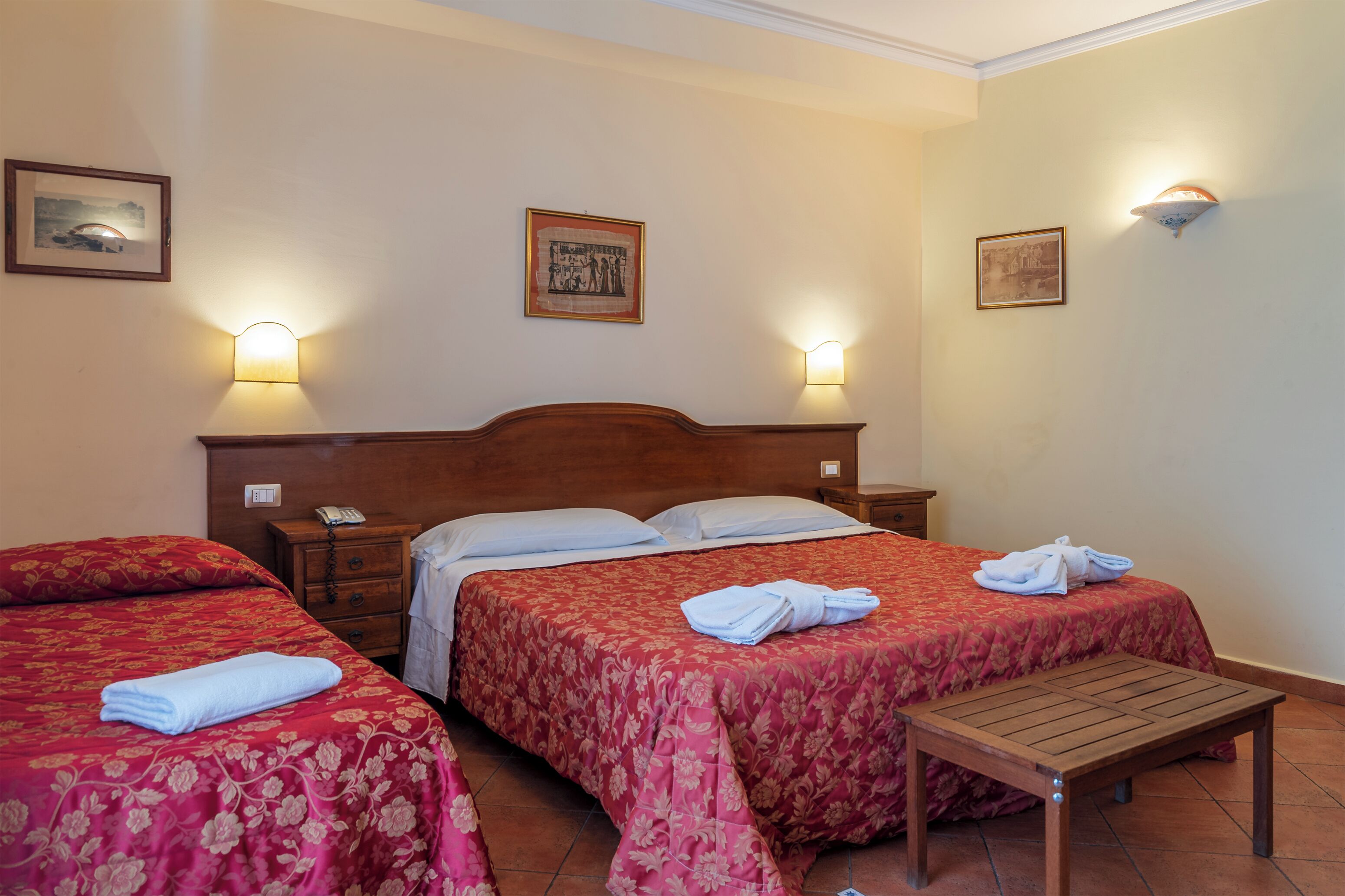 A hotel room with two beds, each with a red floral bedspread, a wooden nightstand, and a small wooden table.