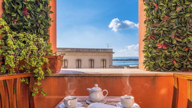 A balcony with a table set for tea, a book, and a view of a building and water. - Hotel Mediterraneo (Syracuse)