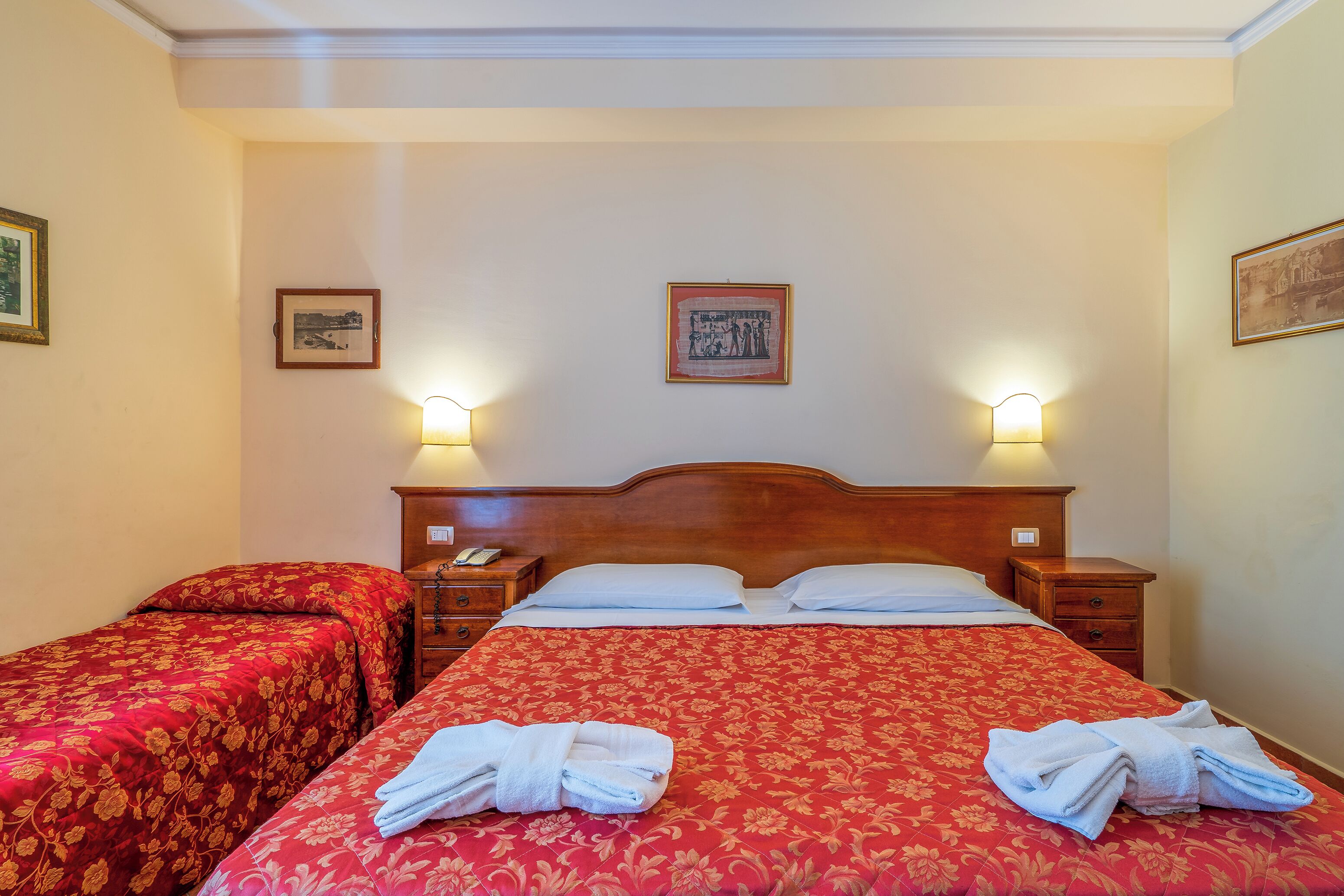 Habitación de hotel con dos camas, cada una con una colcha floral roja y almohadas blancas. Hay dos mesitas de noche con lámparas, y las paredes están decoradas con cuadros enmarcados.