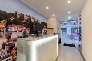 Un área de recepción con un mostrador de vidrio, una televisión montada y un mural de un paisaje urbano con un castillo.