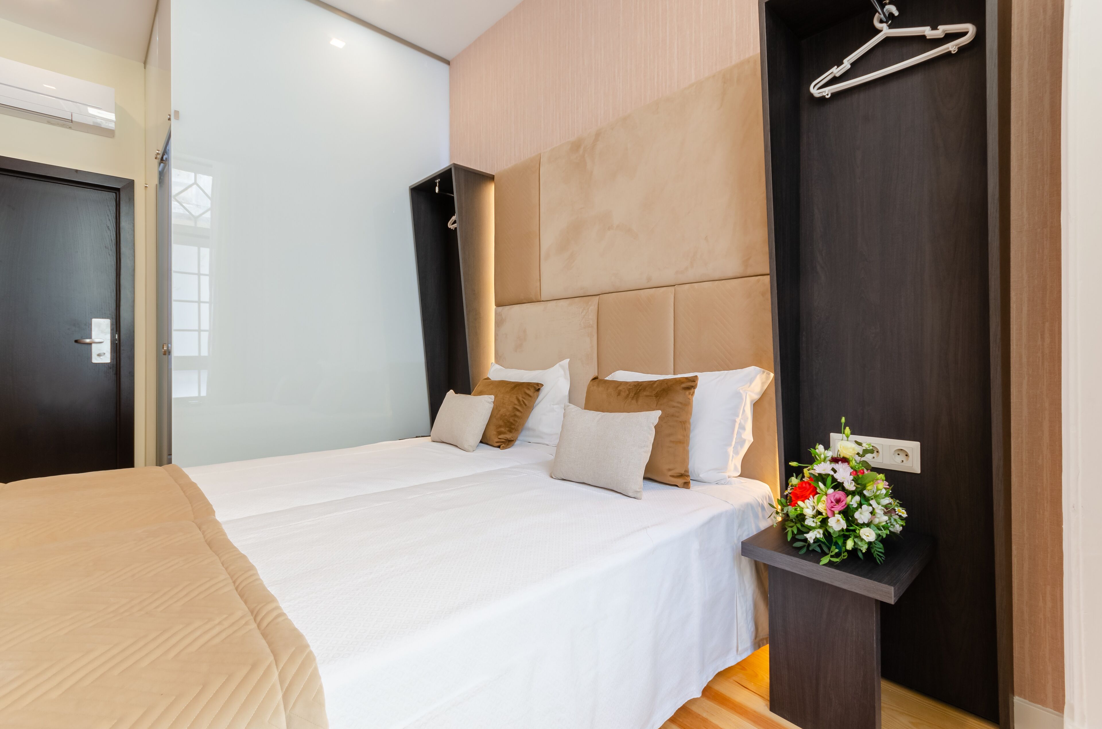 A modern hotel room with a large bed, a bedside table with a flower arrangement, a coat hanger, and a dark headboard.