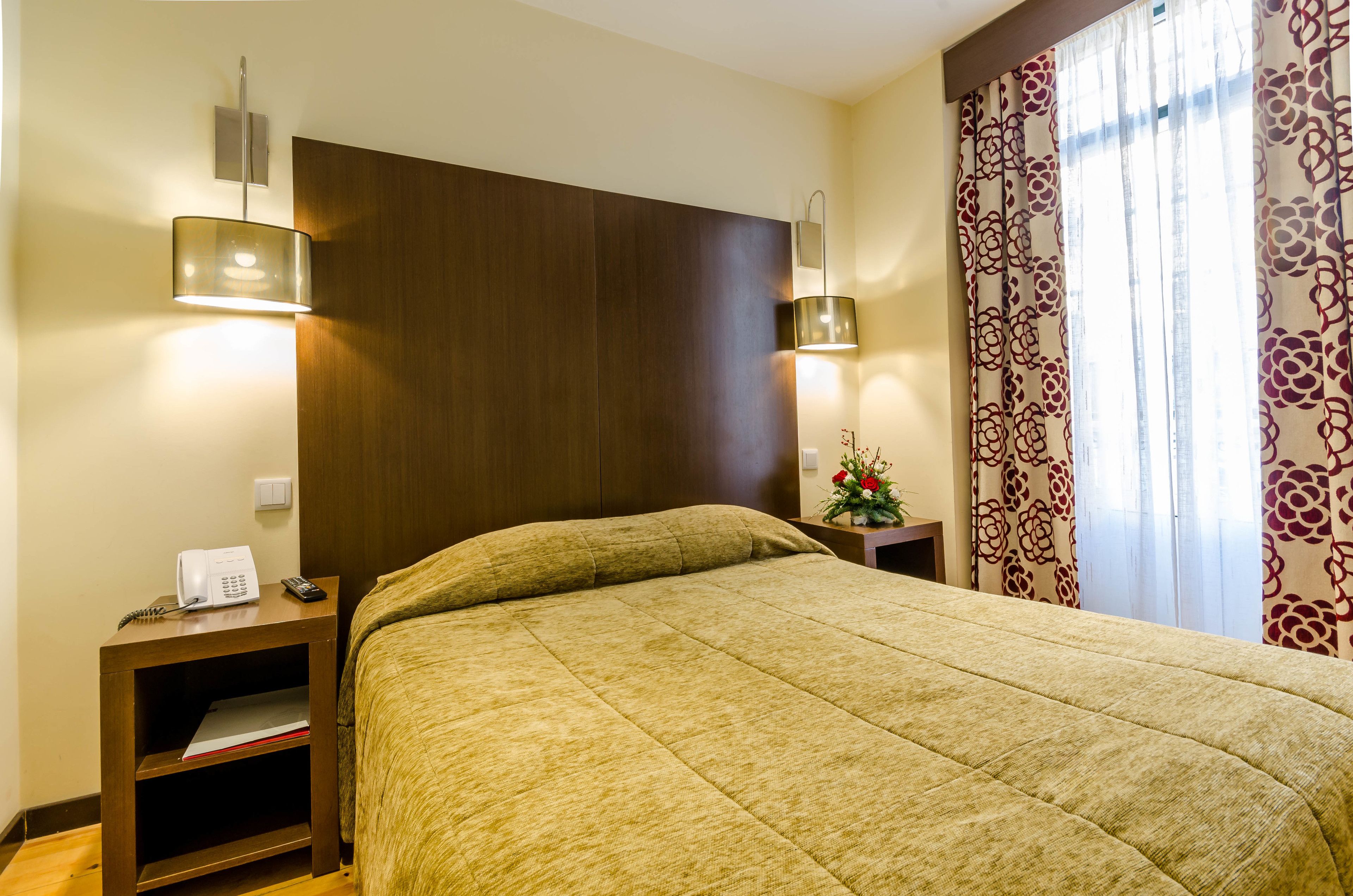 Habitación de hotel con una cama grande, lámparas de noche, mesita con teléfono y ventana con cortinas estampadas.