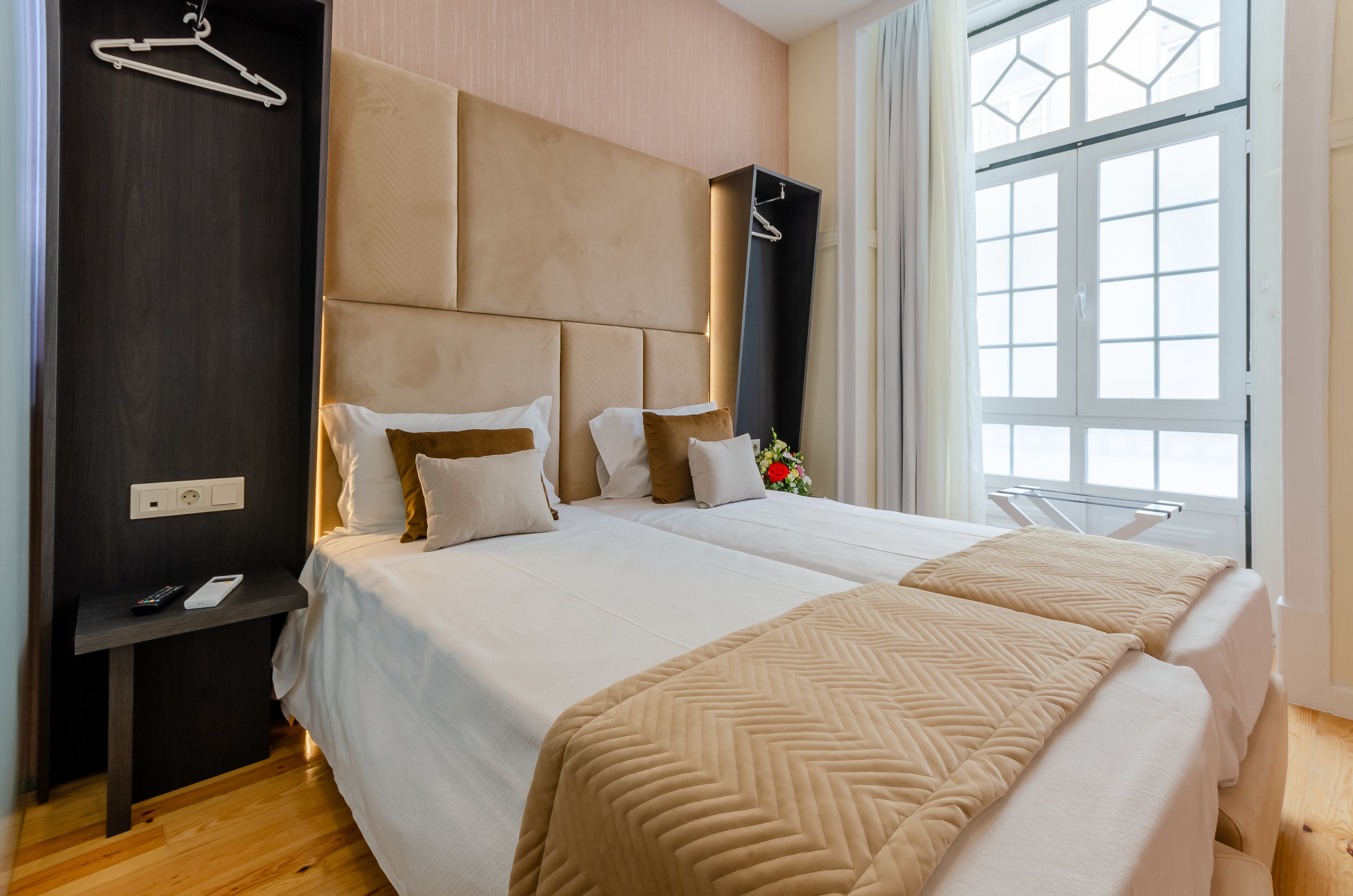 Camera d'albergo con due letti, una testiera in legno scuro, un comodino con telecomando e una finestra con tende.