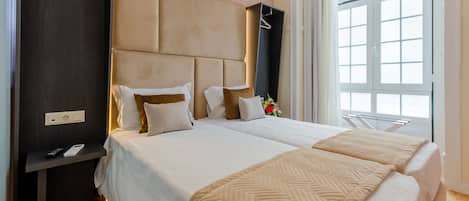 Habitación de hotel con dos camas, cabecero de madera oscura, mesita de noche con un control remoto y ventana con cortinas.