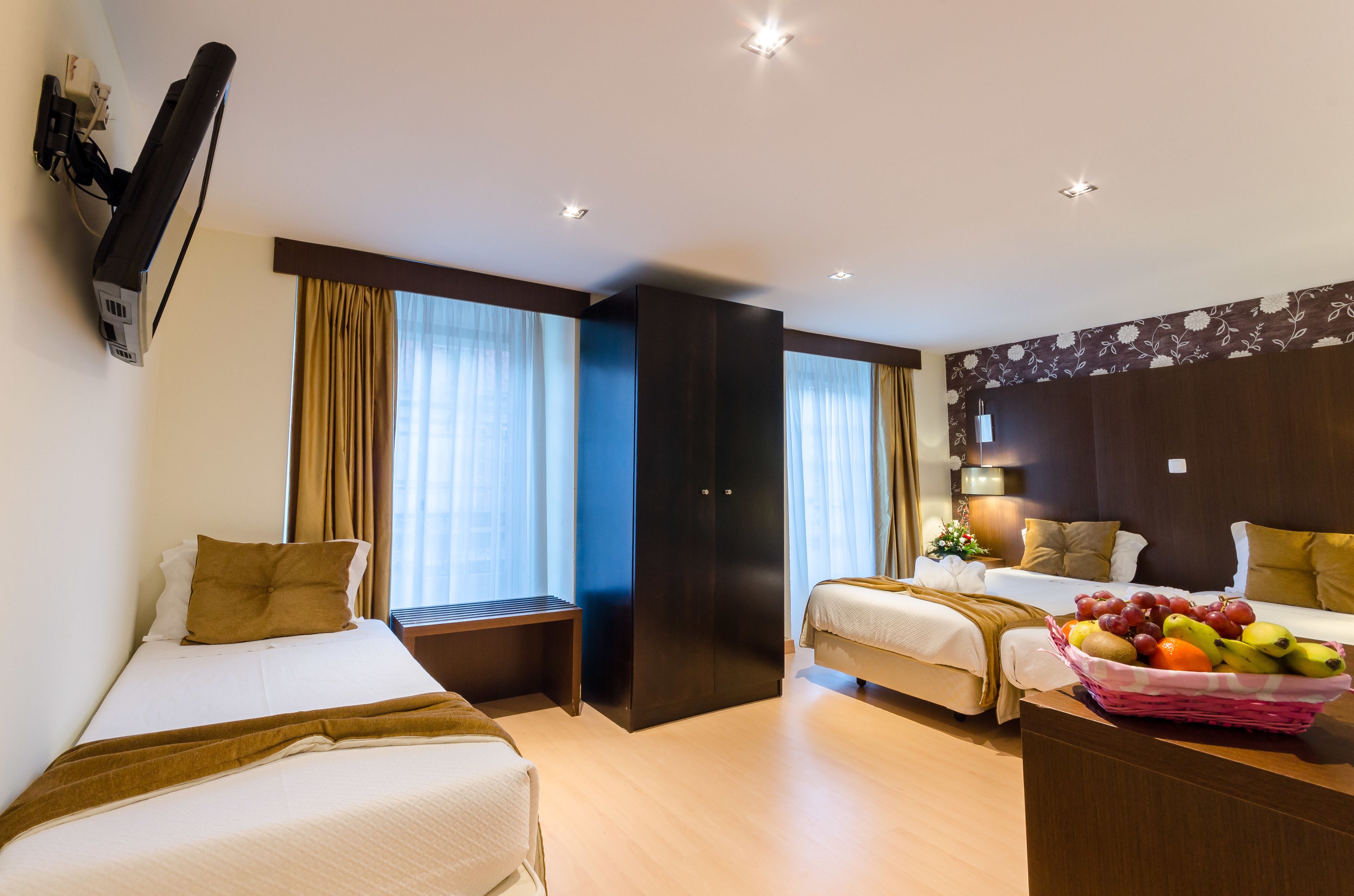 A hotel room with two beds, a desk with a fruit basket, a TV mounted on the wall, and a large wardrobe.