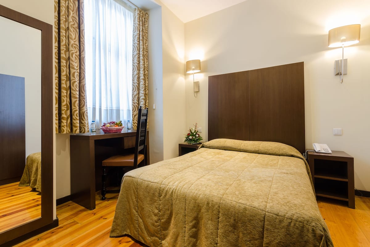 Een hotelkamer met een groot bed, een bureau met stoel, een klein tafeltje met bloemenvaas, een nachtkastje met lamp en een telefoon.