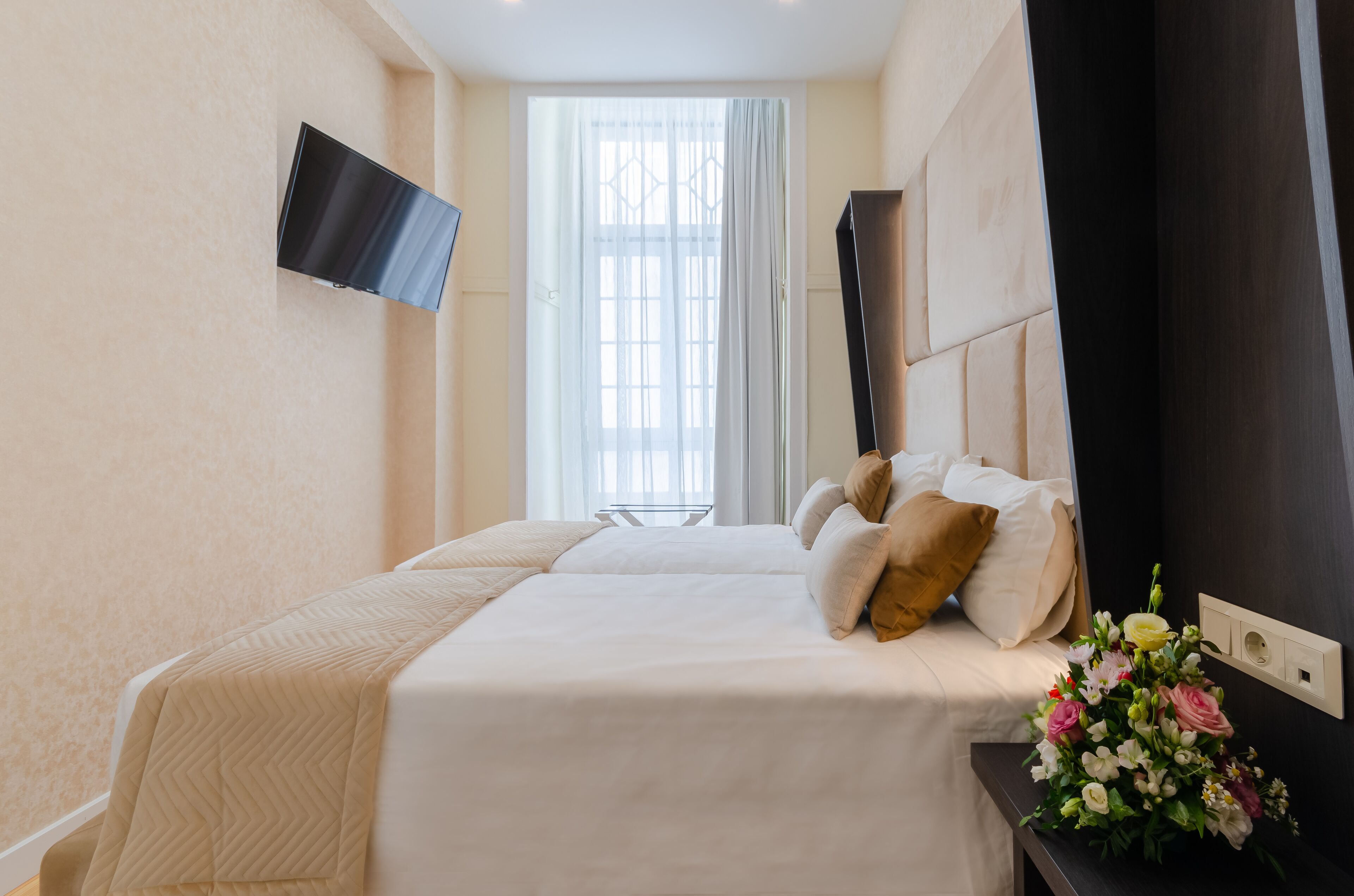 Een hotelkamer met een bed, een aan de muur bevestigde televisie, een raam met gordijnen en een nachtkastje met een bloemenvaas.