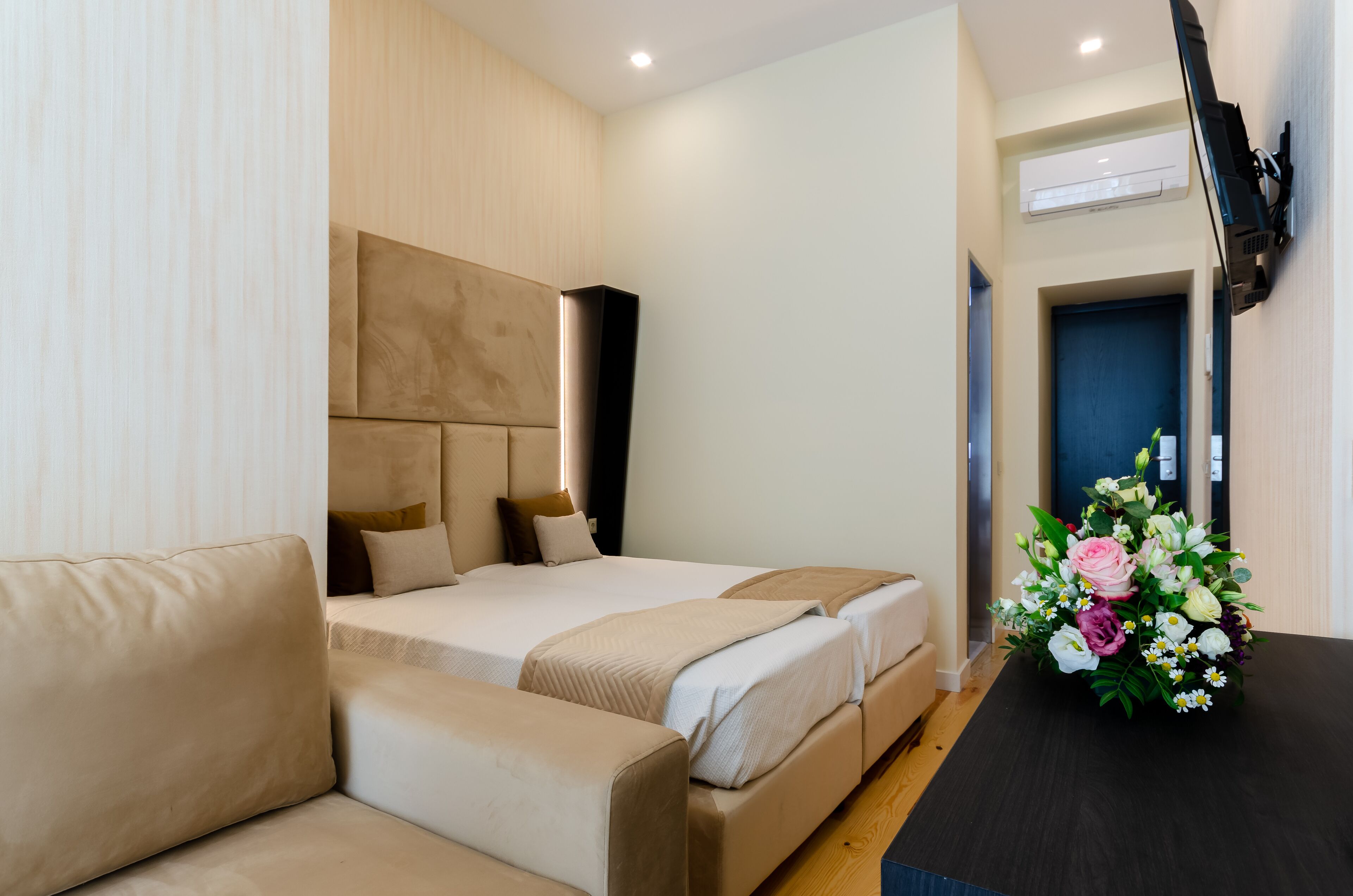 A hotel room with a bed, a sofa, a flat-screen TV mounted on the wall, and a floral arrangement on the table.