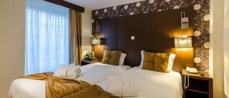 A hotel room with two beds, a nightstand with a lamp, and a floral-patterned wall.