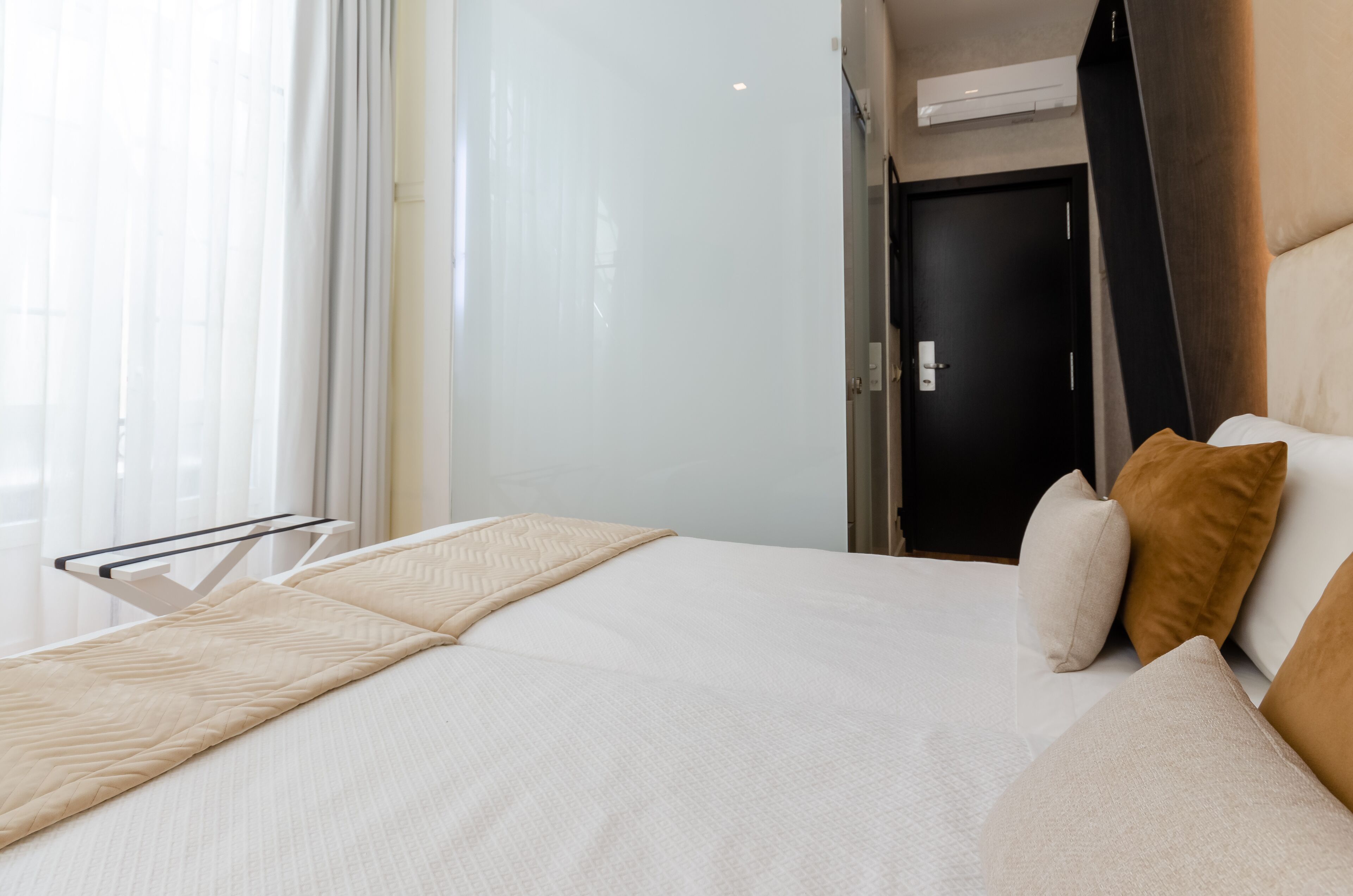 A hotel room with a bed, a window with curtains, a door, and a wall-mounted air conditioning unit.