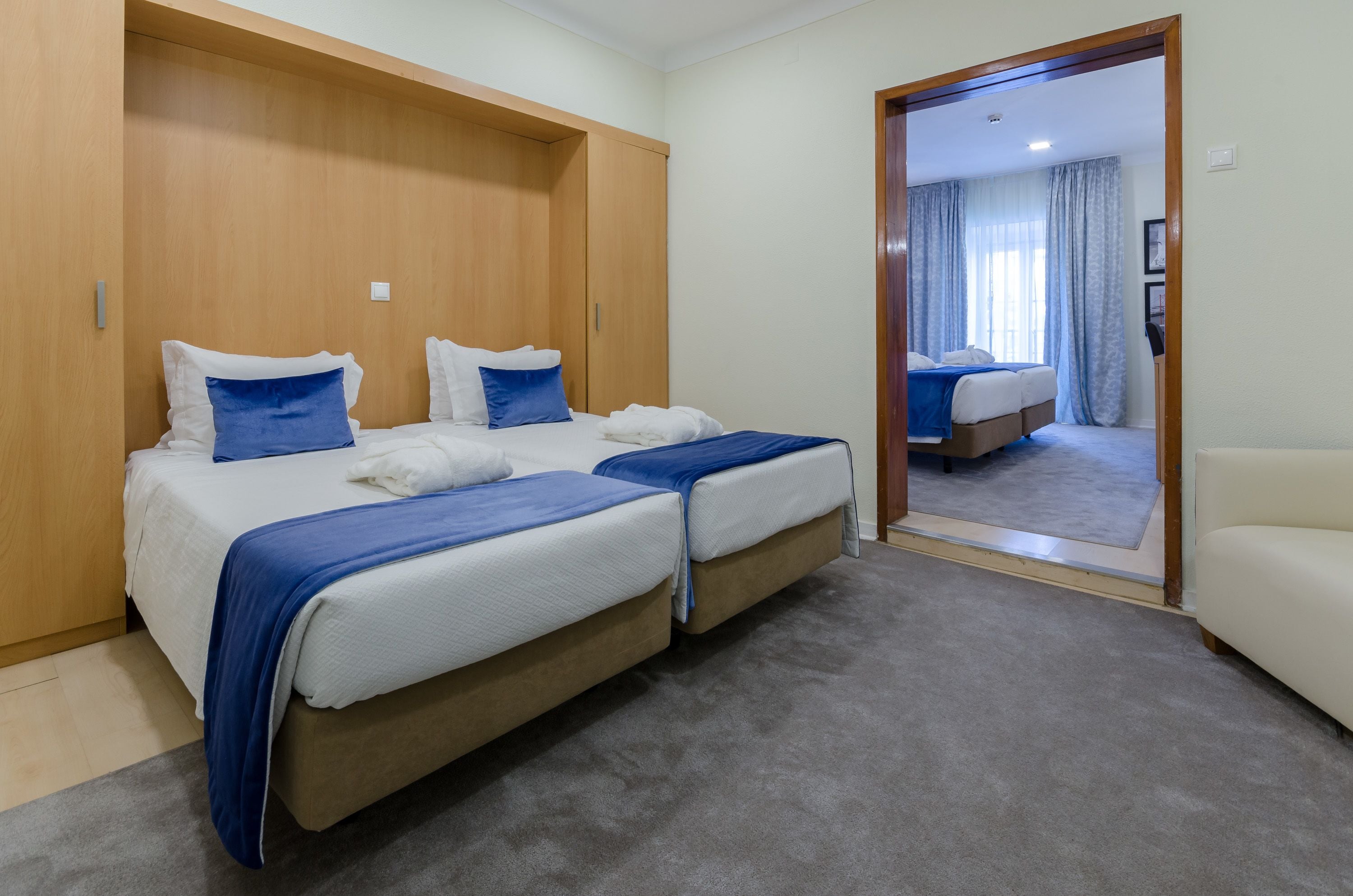 A hotel room with two beds, a wooden wardrobe, a mirror, and a view of another room.