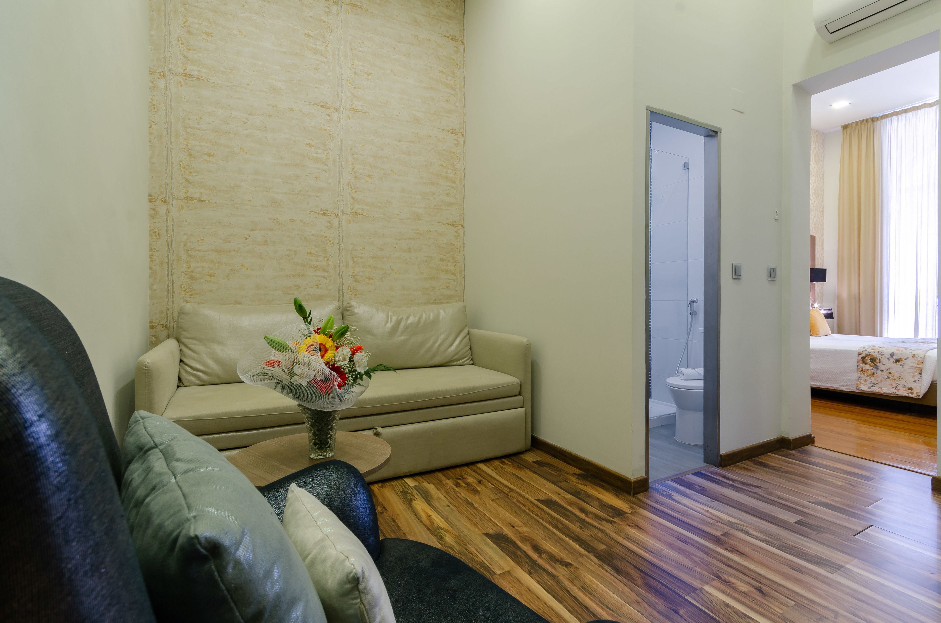 A living room with a beige sofa, a coffee table with a floral vase, wooden flooring, a door leading to a bathroom, and a bed with a floral bedspread.