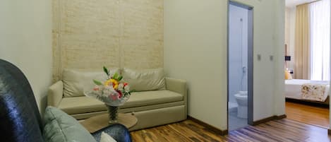 A living room with a beige sofa, a coffee table with a floral vase, wooden flooring, a door leading to a bathroom, and a bed with a floral bedspread.
