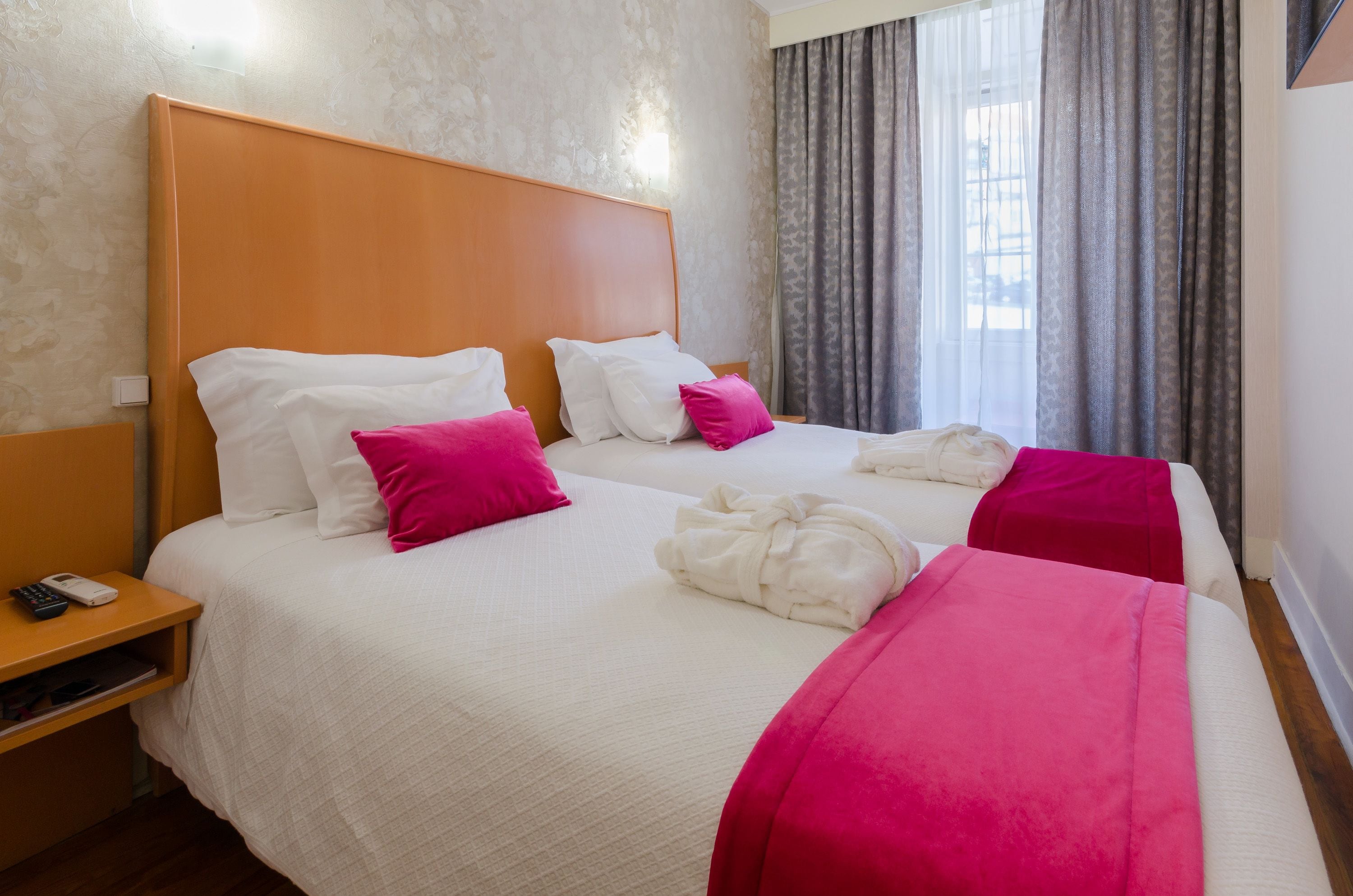 A hotel room with two beds, each with white bedding and pink accents, a wooden headboard, and a nightstand with a phone.