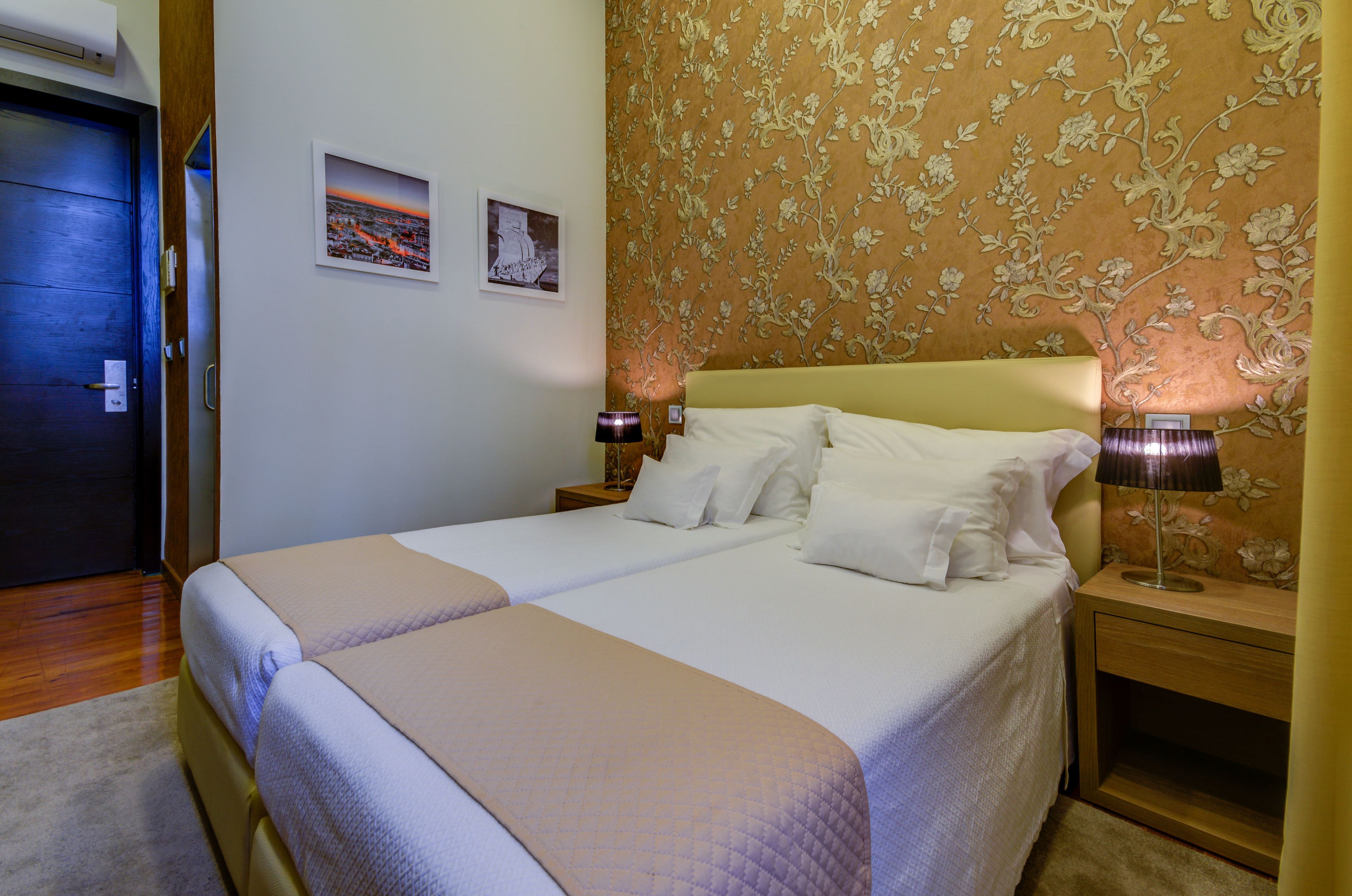 A double bed with a beige headboard, two bedside tables with lamps, and a floral wallpaper pattern on the wall.