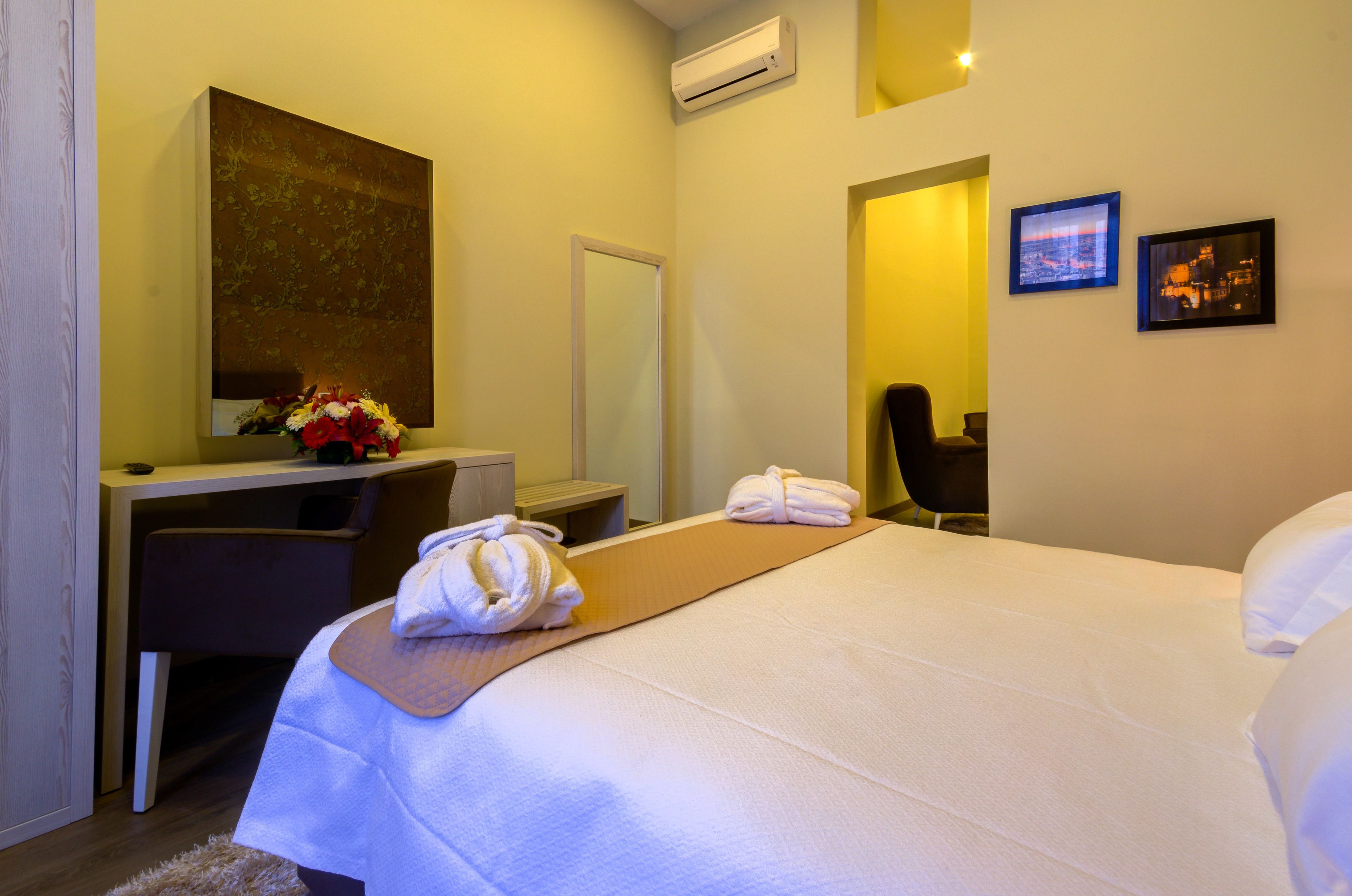 A hotel room with a bed, a desk, a chair, a mirror, and two framed pictures on the wall.