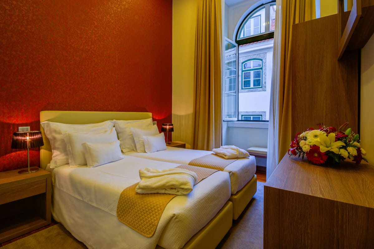 A hotel room with two beds, a wooden nightstand, a bedside table with a lamp, a wooden dresser, and a window with curtains.