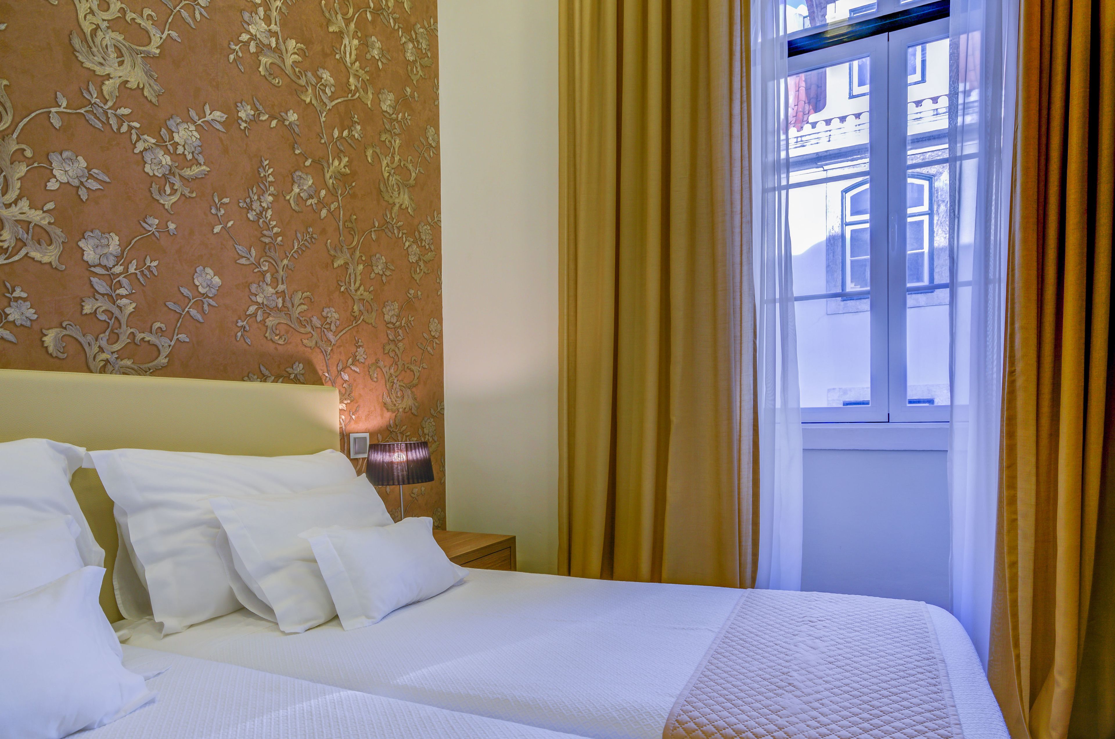 A hotel room with a bed, pillows, a nightstand, a lamp, and a window with curtains.
