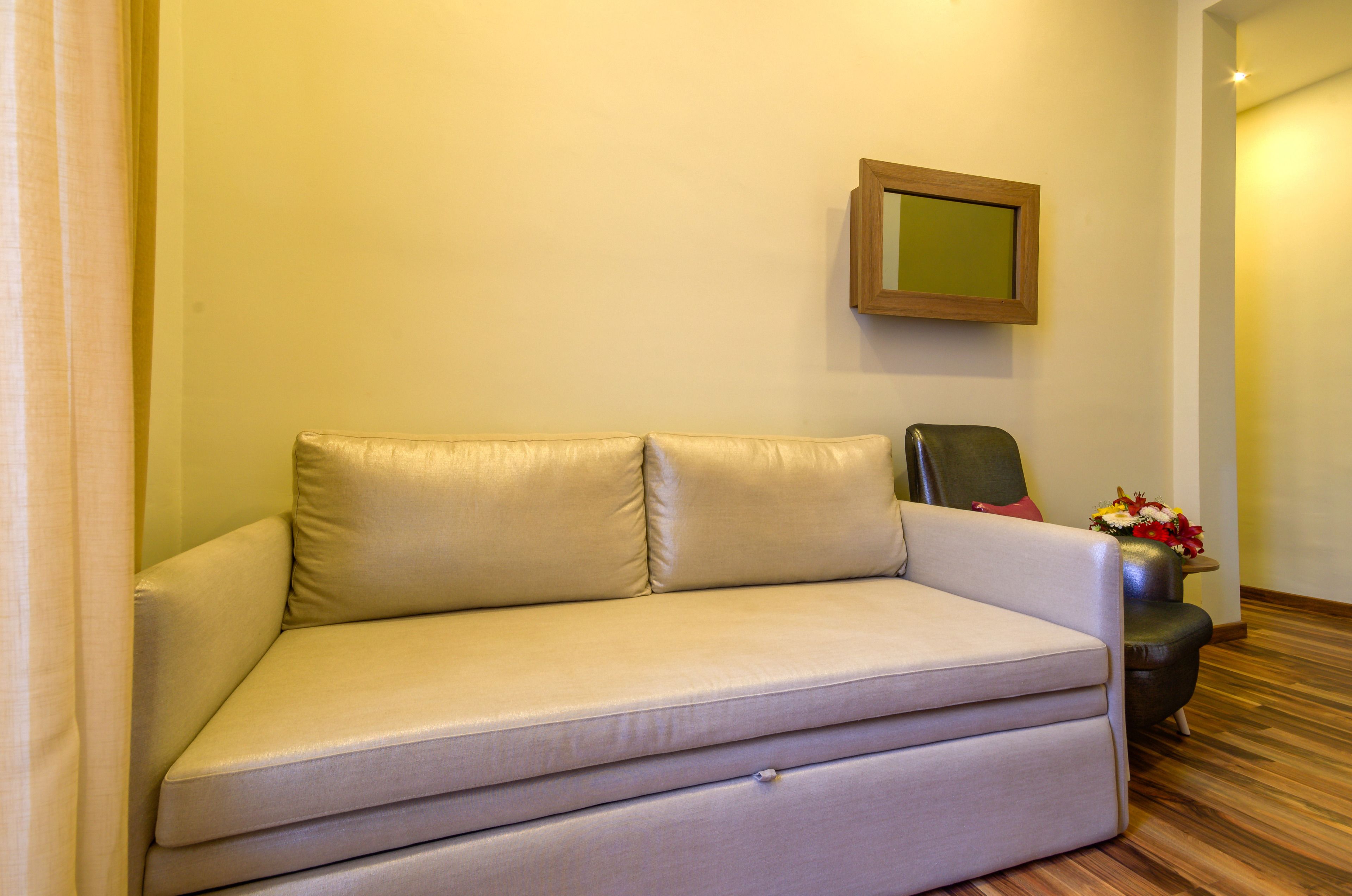 Een beige bank in een kamer met een houten vloer, een klein tafeltje met een bloevas en een lijst met spiegel aan de muur.