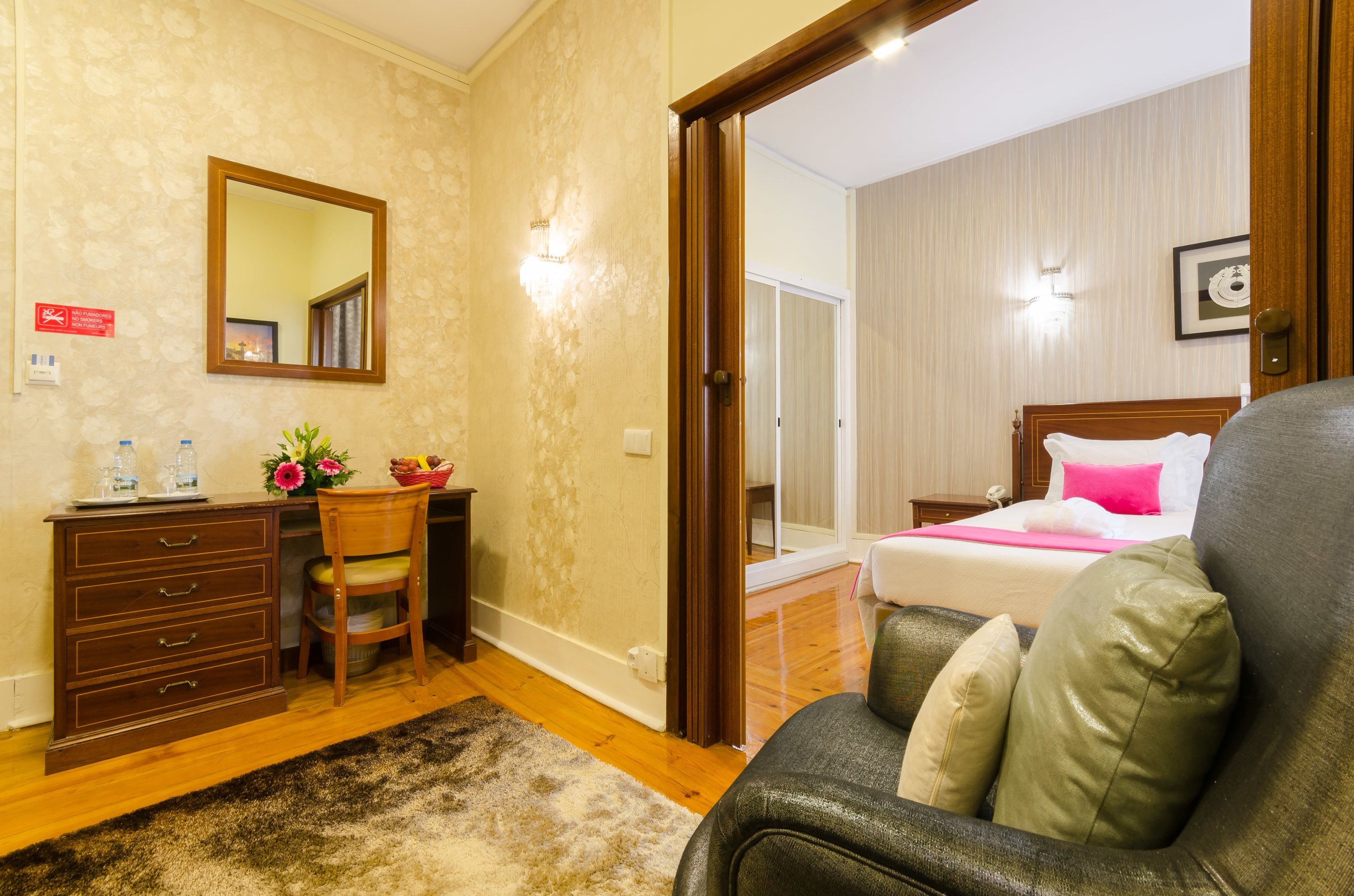 A hotel room with a wooden dresser, a chair, a bed with a pink pillow, and a mirror on the wall.