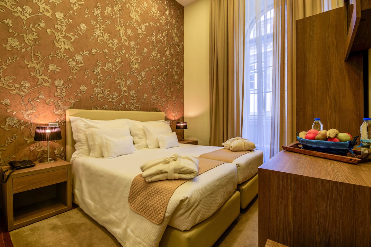 A hotel room with a large bed, a bedside table with a lamp, a nightstand, a wooden dresser, and a window with curtains.