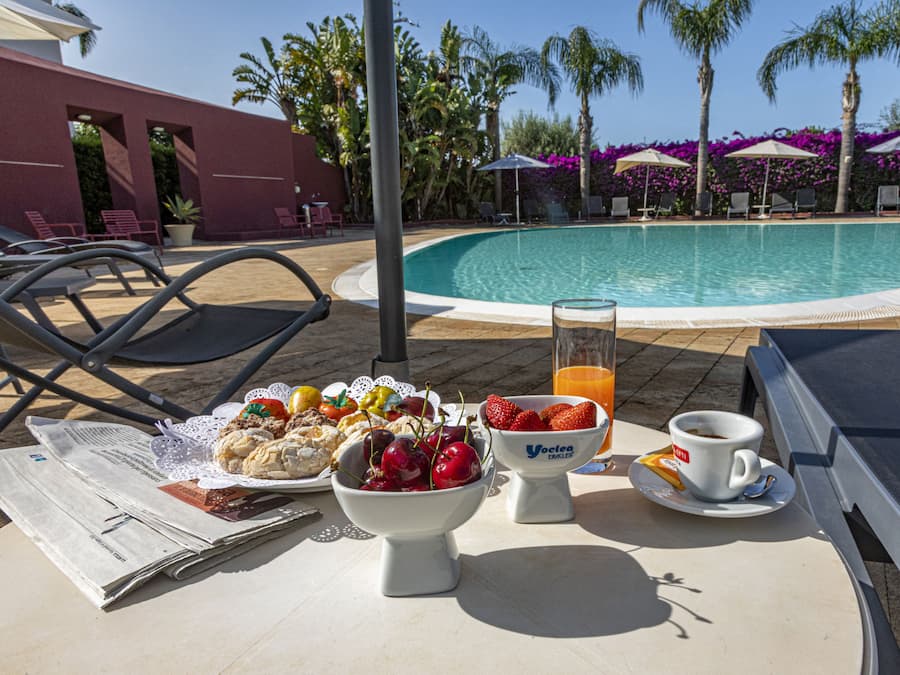 A poolside breakfast setup with a newspaper, fruit bowl, juice, and coffee.